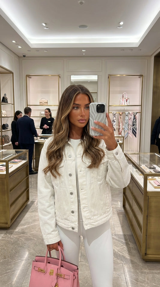 Trendy woman holding a phone with a gray and silver zebra patterned case, wearing white attire and carrying a pink handbag in an elegant boutique setting.