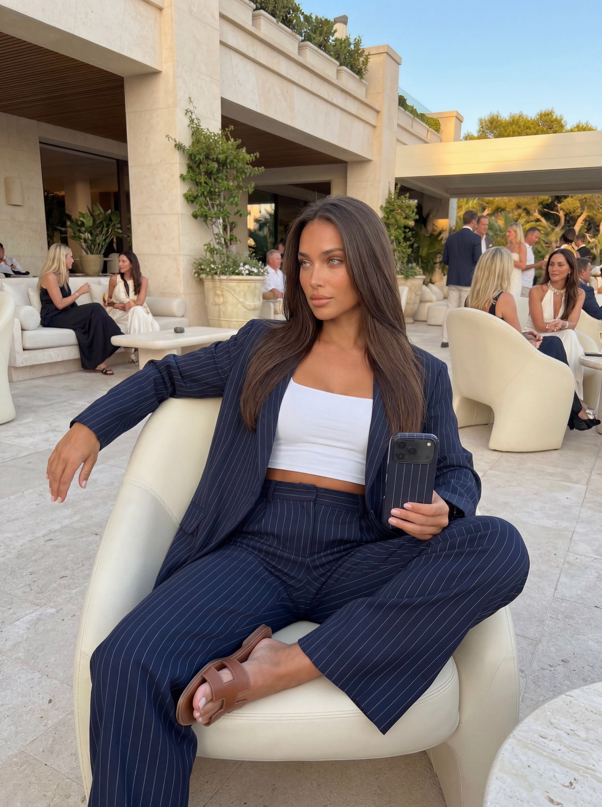 Trendy woman wearing a blue and black pinstripe suit with a white crop top, sitting confidently while holding a phone case in an elegant outdoor setting.