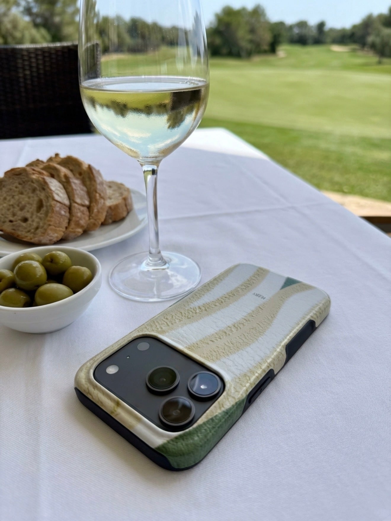 Tricolore Matcha phone case with grøn, hvid, and flerfarvet patterns beside a glass of white wine, olives, and bread on a table with a scenic outdoor view.