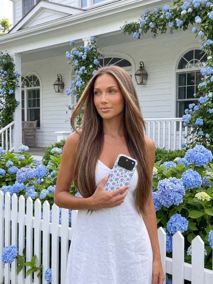 Trendy woman in white dress holding a phone case with a multicolored blue and white floral pattern, standing by a white picket fence and blooming hydrangeas.